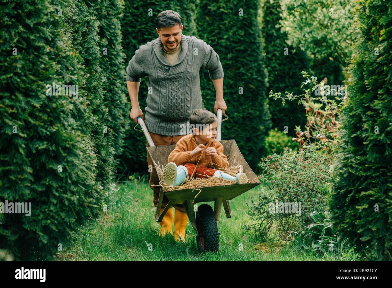 Father pushing son sitting on hay in wheelbarrow at back yard Stock Photo