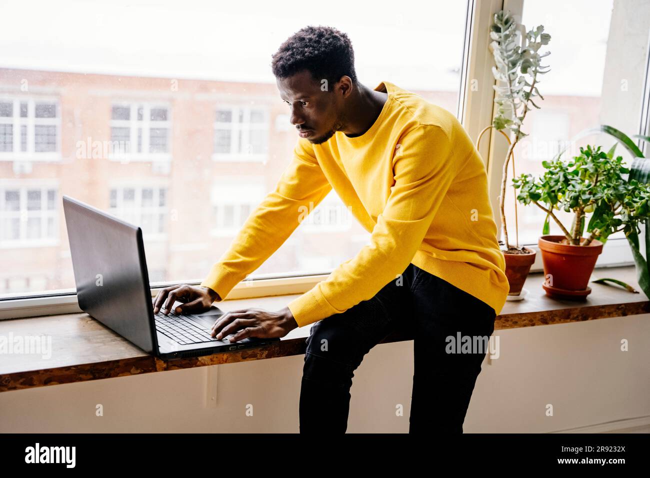 Focused man using laptop sitting on window sill at home Stock Photo