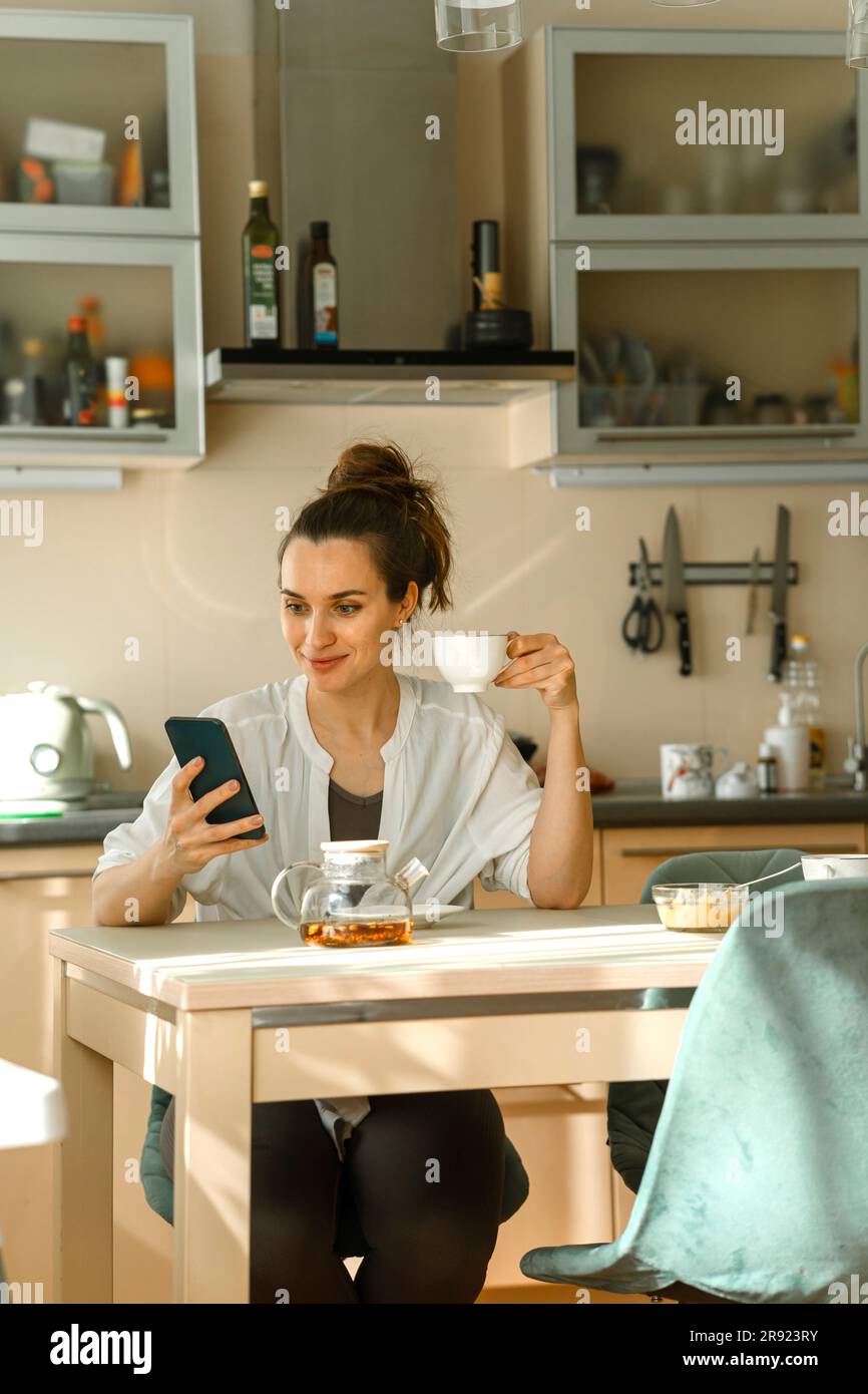 Smiling woman using smart phone and having tea at home Stock Photo
