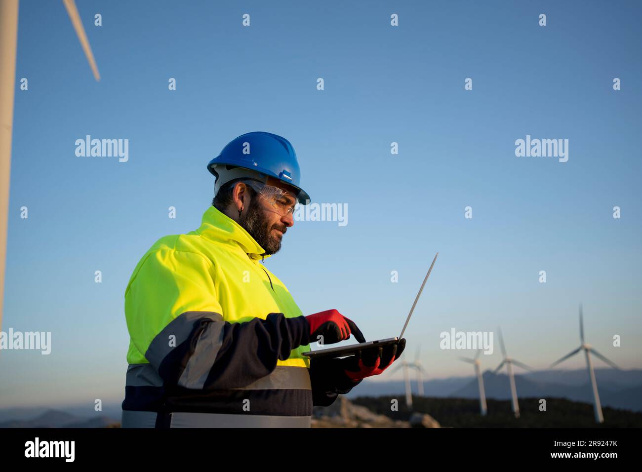Engineer wearing hardhat using laptop near wind turbines Stock Photo