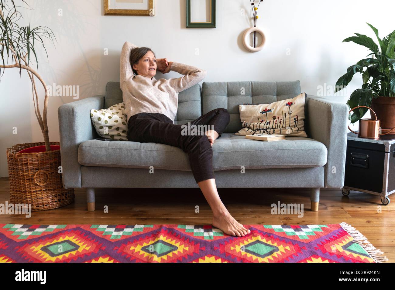 Thoughtful mature woman with hands behind head sitting on sofa at home ...
