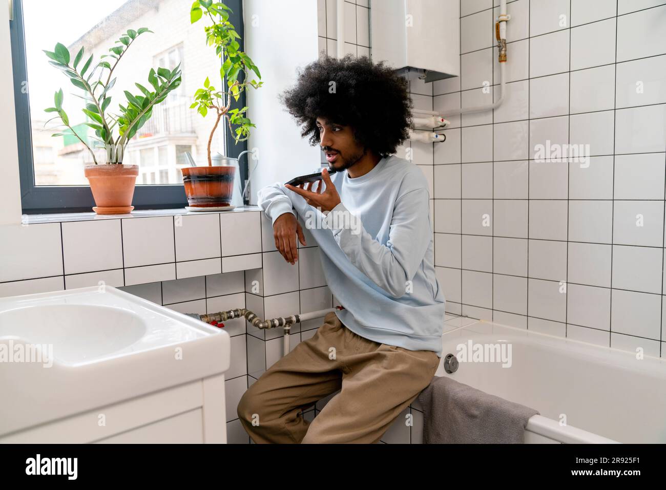 Man talking on speaker phone sitting on bathtub in bathroom at home Stock Photo