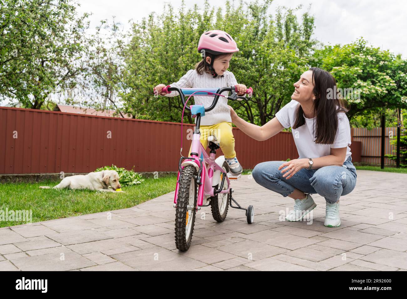 Smiling mother crouching by daughter learning to ride a bicycle on footpath Stock Photo