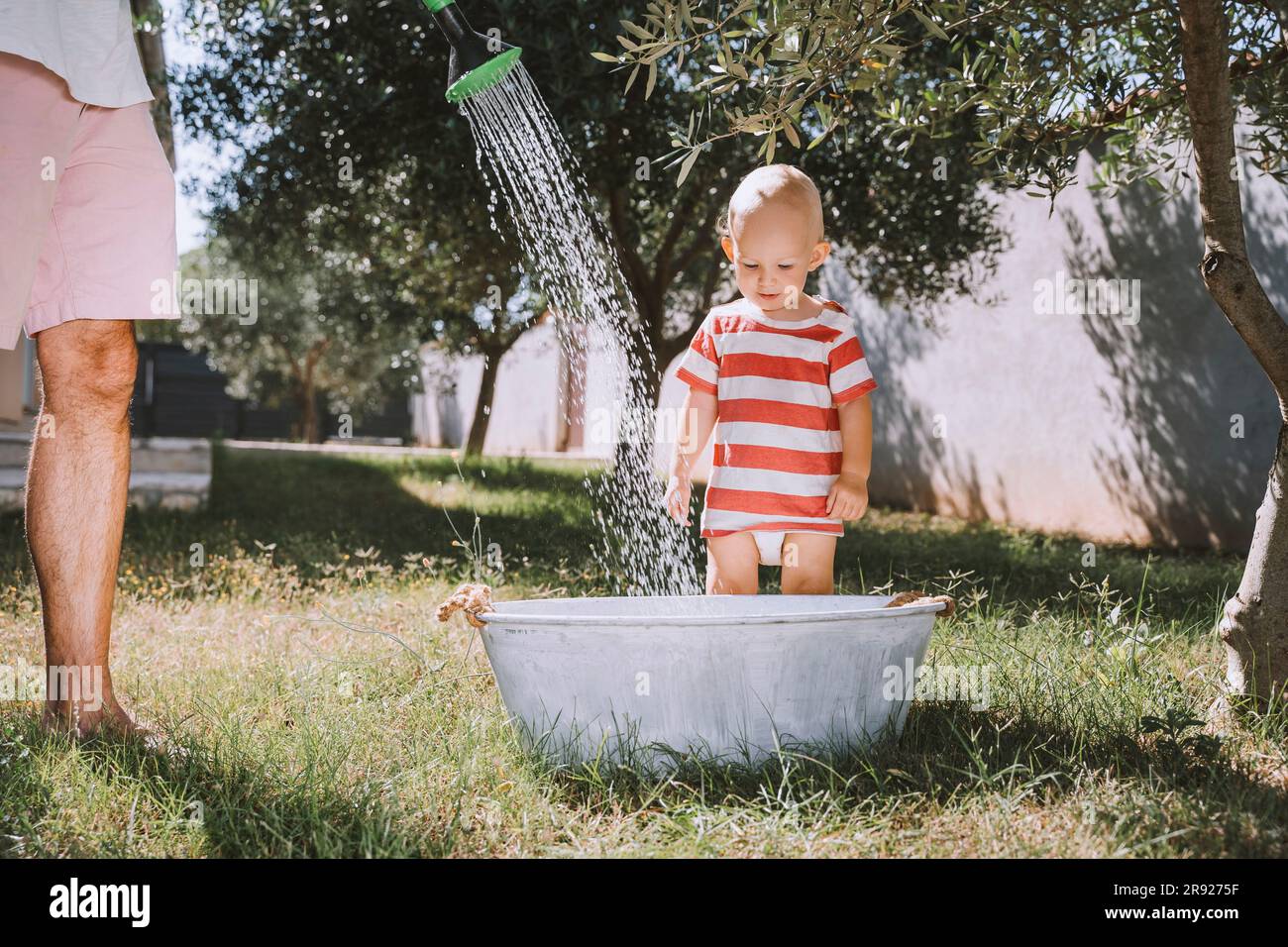 Toddler standing with father pouring water in bathtub at backyard Stock