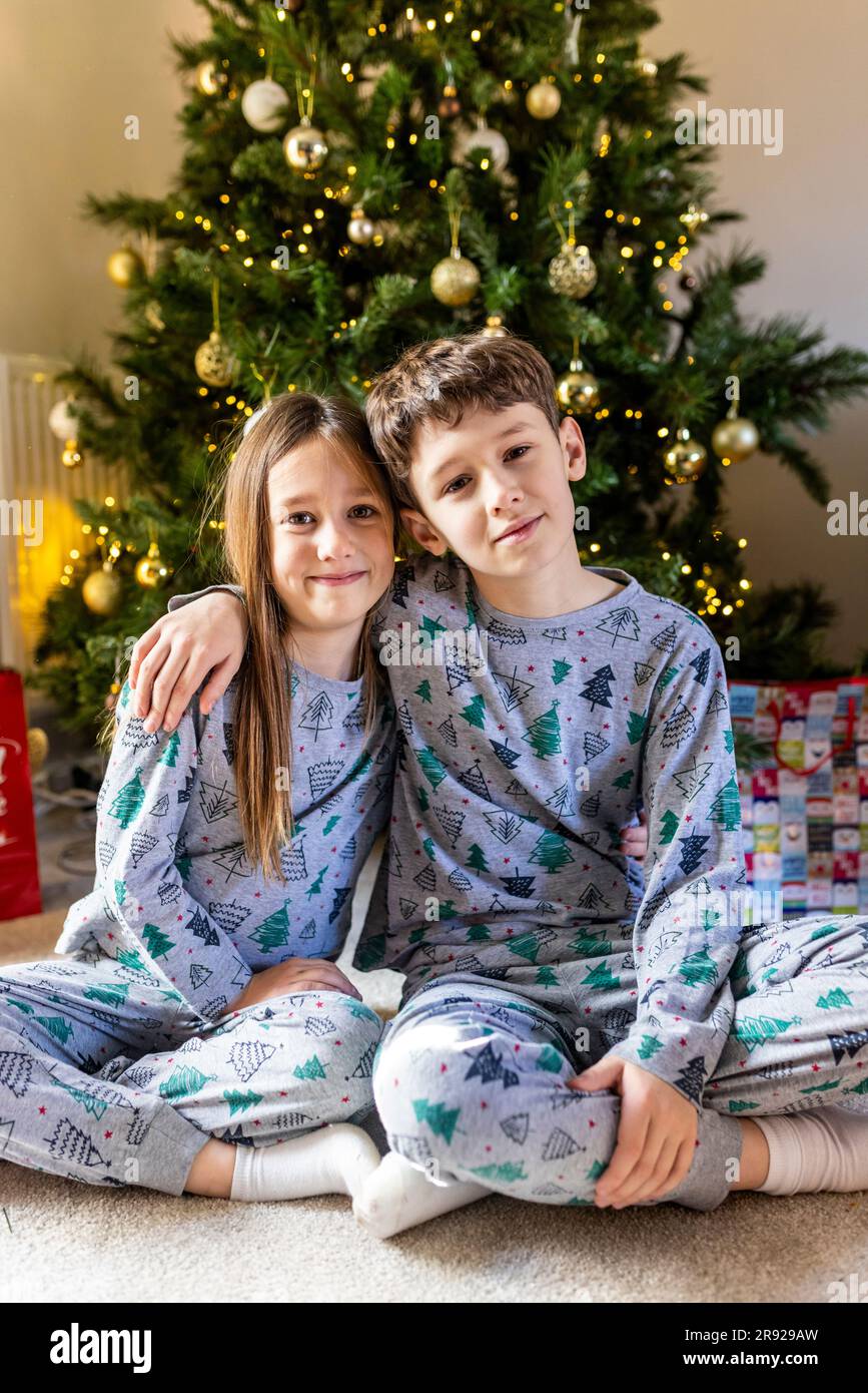 Smiling brother and sister sitting in front of Christmas tree Stock Photo