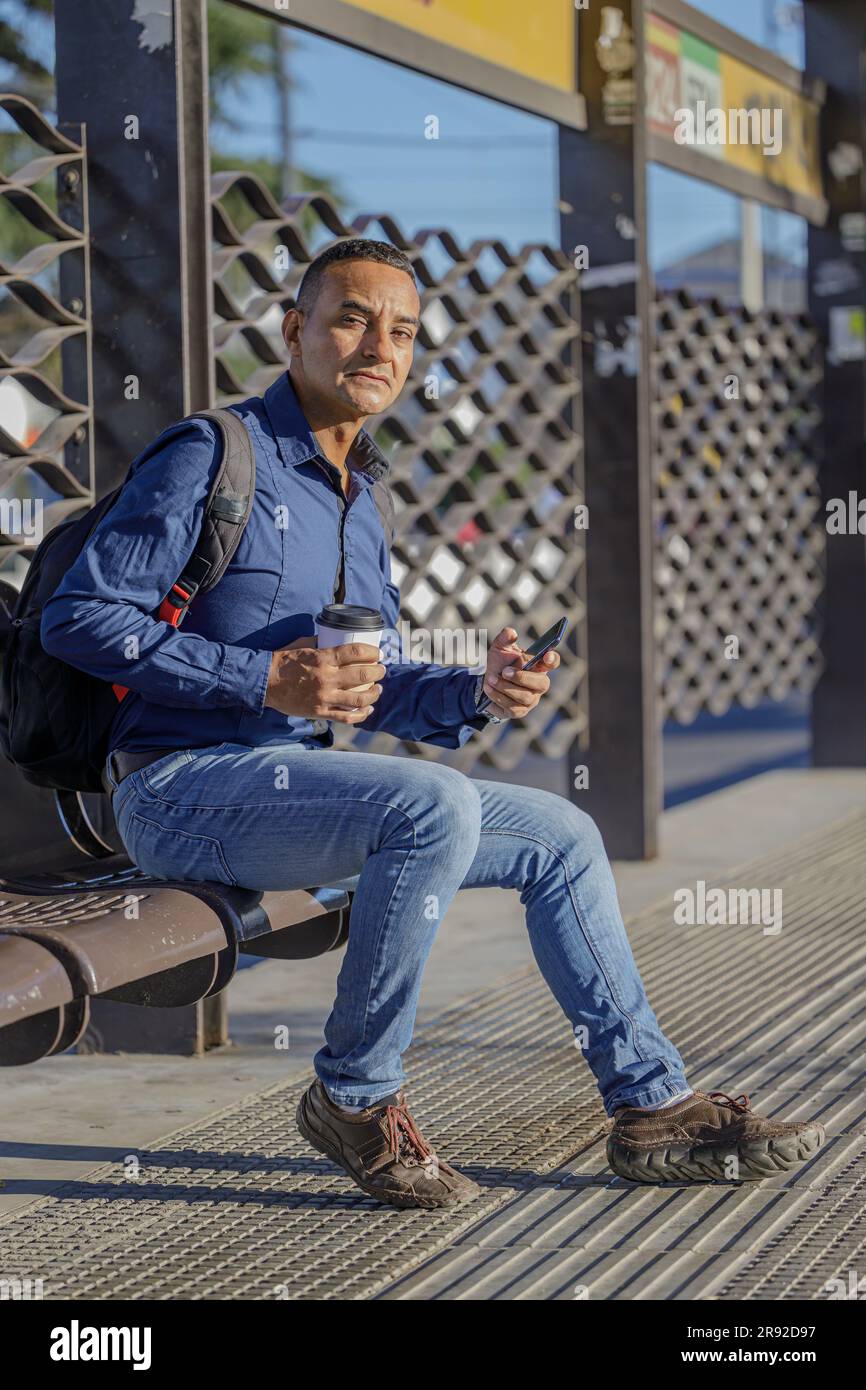 Young latin man sitting waiting for the bus with a paper cup of coffee in his hand. Stock Photo