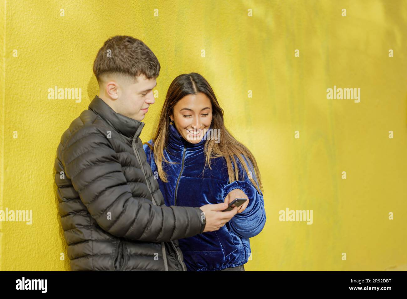 Young couple look at a mobile phone with a yellow wall in the background with copy space. Stock Photo