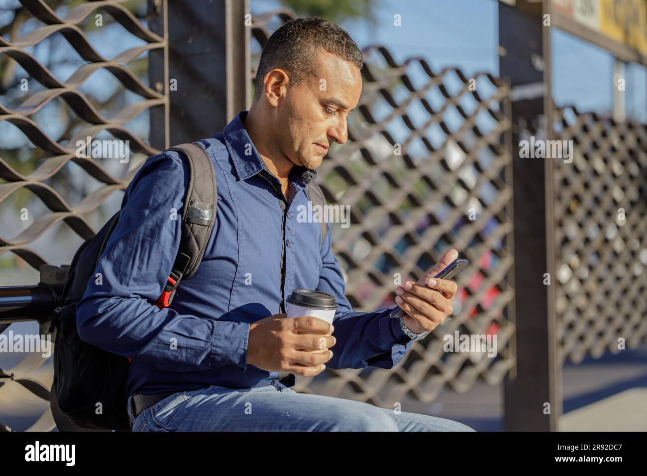 Young latin man sitting looking his mobile phone and waiting for the bus with a paper cup of coffee in his hand. Stock Photo