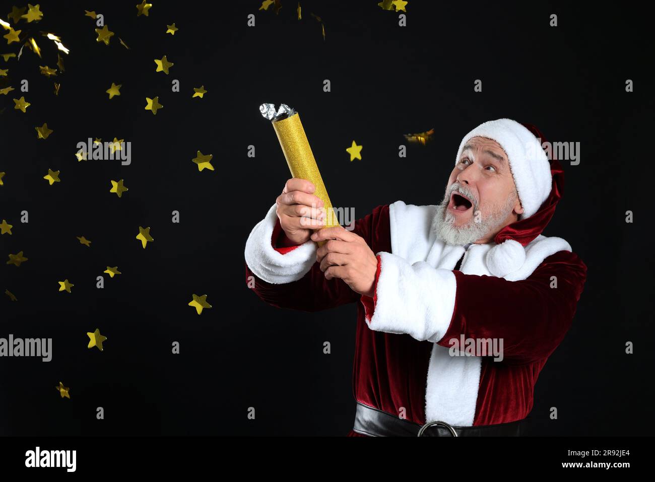 Emotional man in Santa Claus costume blowing up party popper on black background Stock Photo