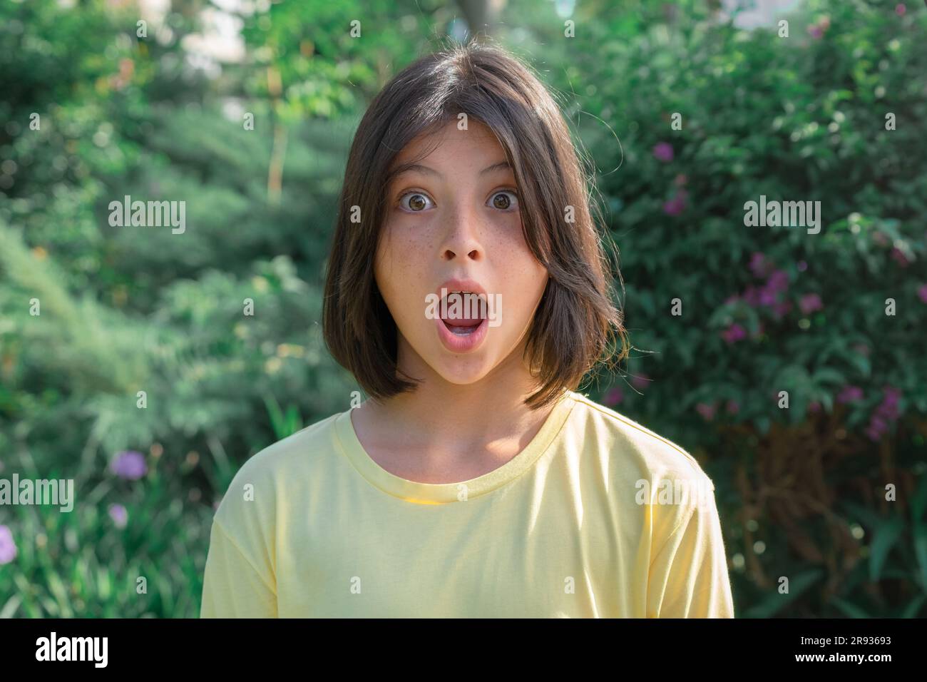 Close-up portrait of a very surprised teenage girl with wide open eyes and mouth against the background of greenery. Stock Photo