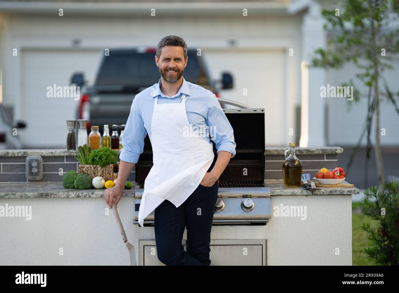 glad cook man at bbq and grill hold utensil. cook man at bbq and grill outdoor. photo of cook man at bbq and grill in apron. cook man at bbq and grill Stock Photo