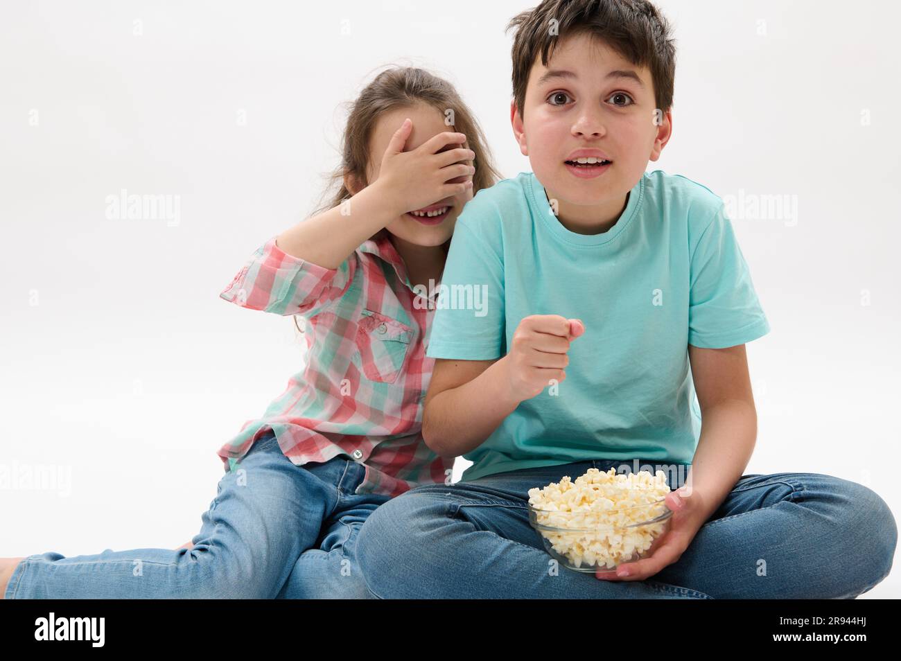 Amazed kids, teen boy and little child girl watching scary movie or cartoon, eating popcorn on white isolated background Stock Photo
