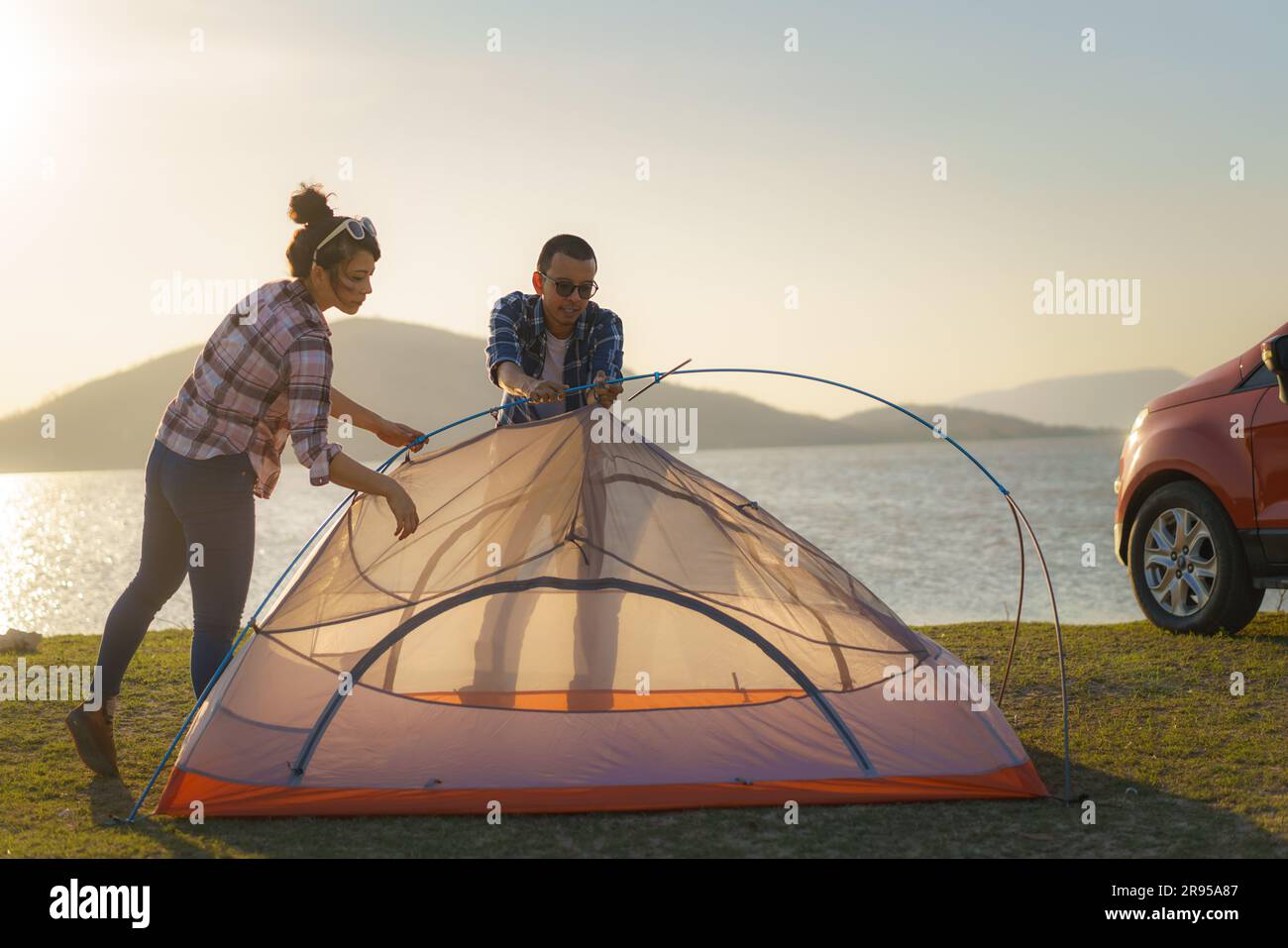 Asian couple preparing a tent to camping in the lawn with the lake in the background during sunset Stock Photo