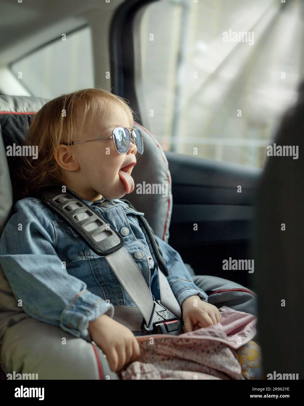 A baby in his mother's sunglasses sits in a car in a child seat and shows his tongue out the window Stock Photo