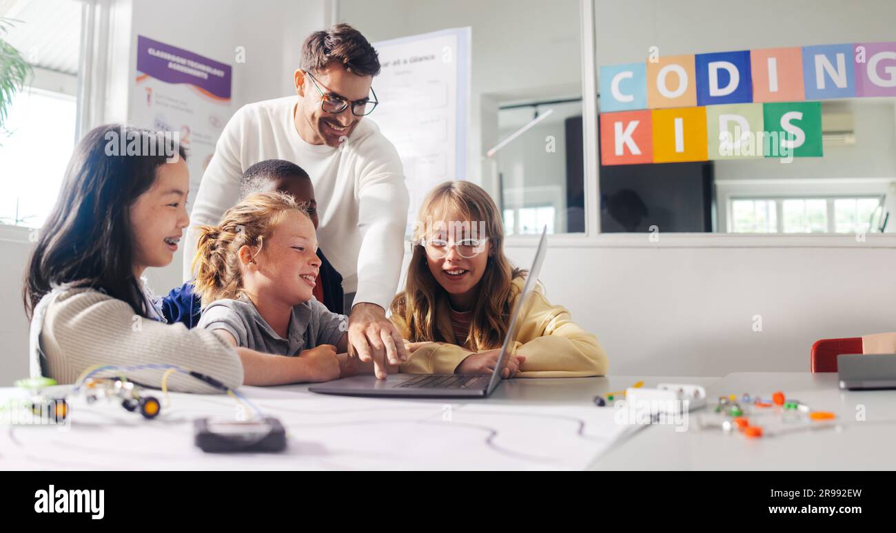 Male teacher guides elementary school students in a robot programming class. Kids learn to code using education software on a laptop and control robot Stock Photo