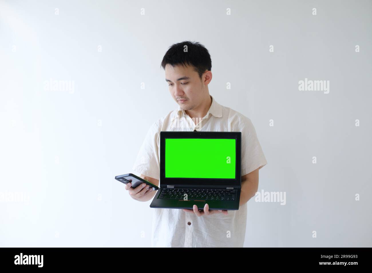 An Asian man wearing a beige shirt is using and looking at a smartphone while holding a laptop with green screen. Isolated white background. Stock Photo