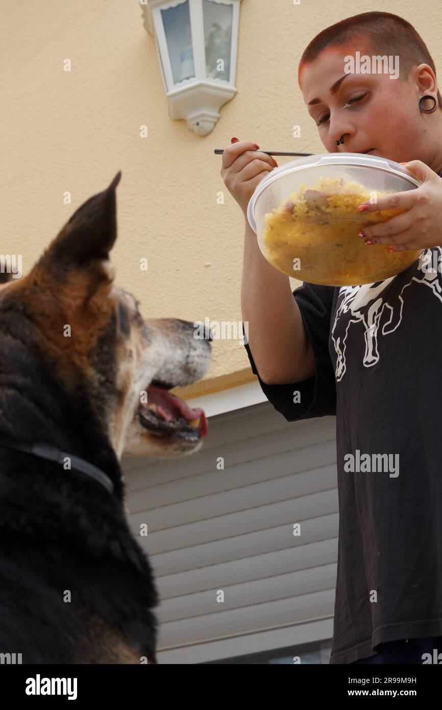 Young woman eating from grandmother's rice tupperware while her dog watches and waits for some rice to fall out Stock Photo