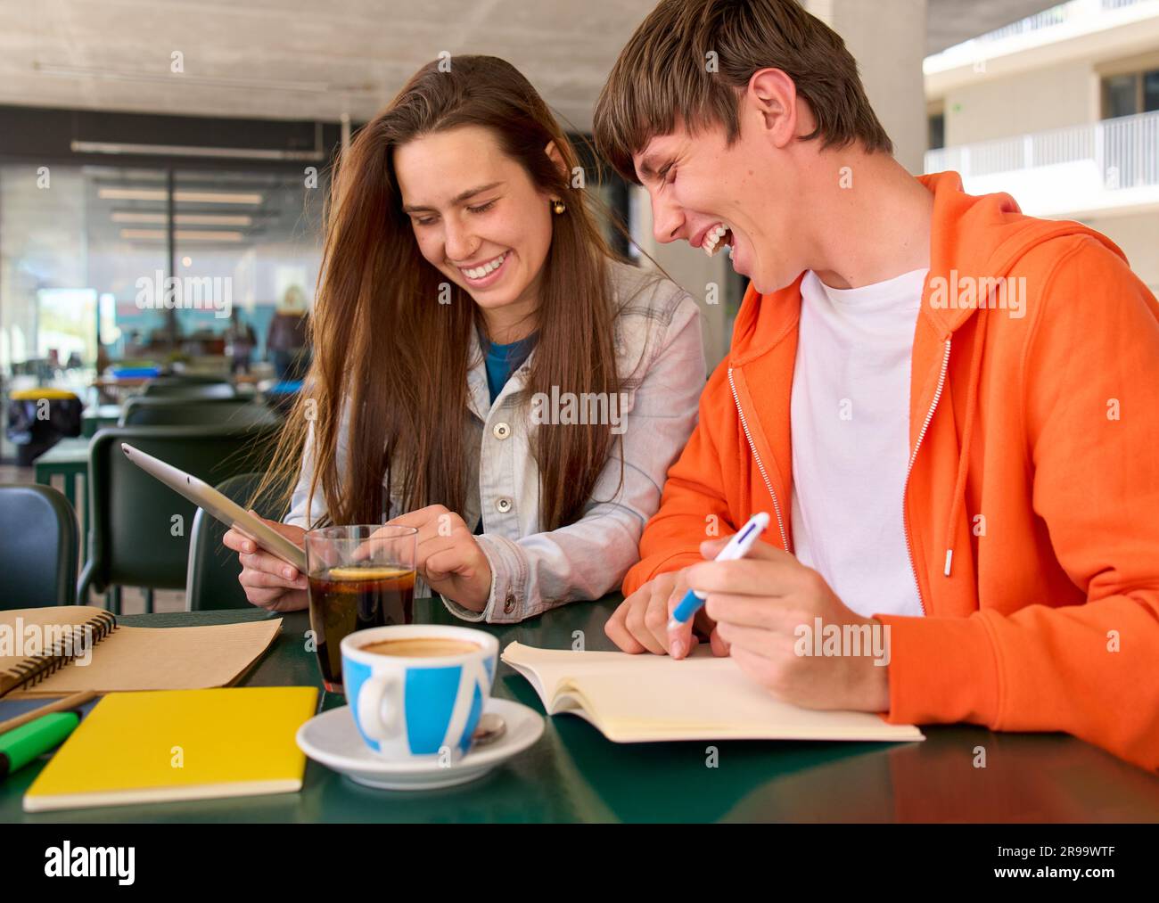 Young Caucasian female and male laughing while using tablet notepad in university campus cafeteria.  Stock Photo