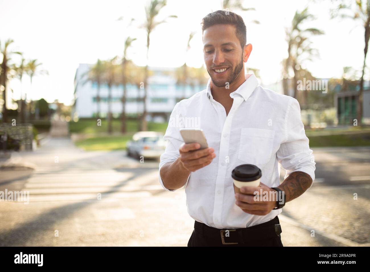 Happy businessman using smartphone and having coffee while walking outdoors during break, free space Stock Photo