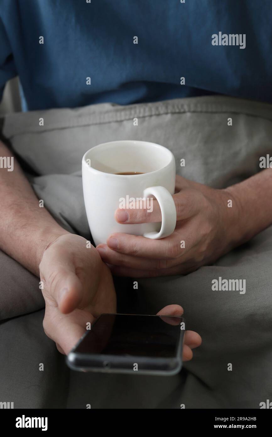 hands of man with cell phone and cup of coffee in bed Stock Photo