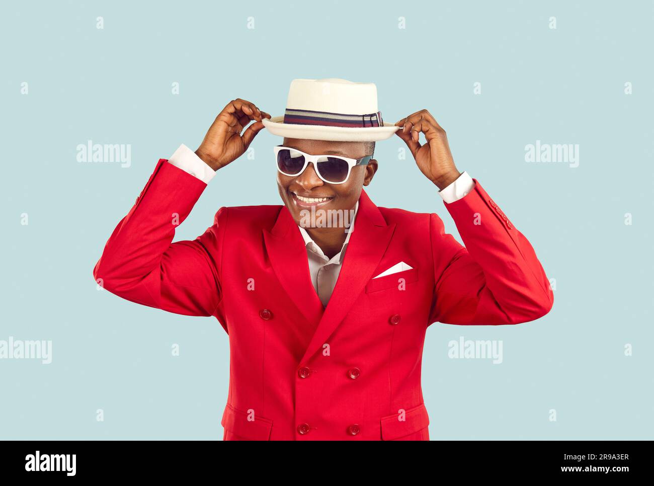 Funny smiling dark skinned man in stylish red jacket adjusts his hat on pastel light blue background Stock Photo
