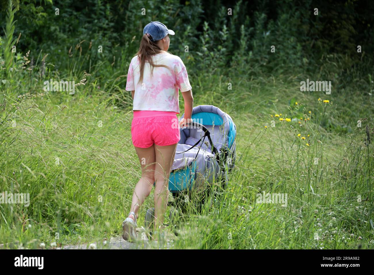 Girl in pink shorts walking with baby pram in a park. Young mother, leisure at summer nature Stock Photo