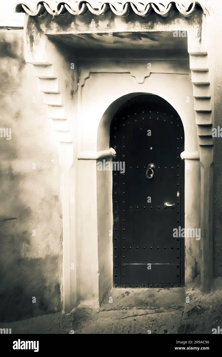 An aged rendering of an An exterior wall and a formidable arched wooden door, framed with architectural details  Chefchaouen, Morocco Stock Photo