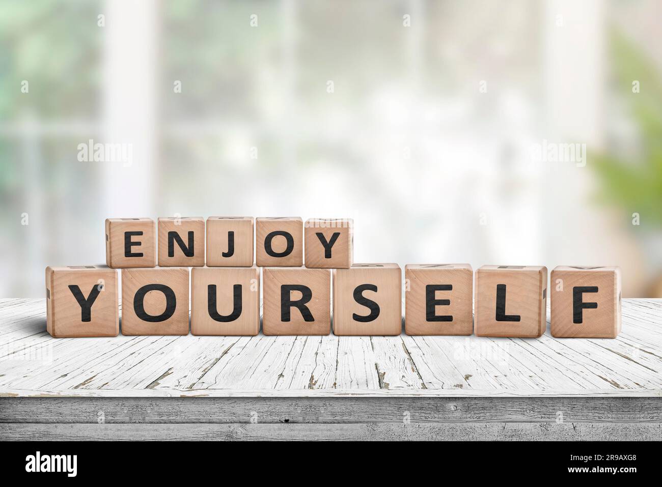 Enjoy yourself wriiten on wooden blocks in a bright room. Cubes with a positive phrase on a white desk in a living room with green plants Stock Photo