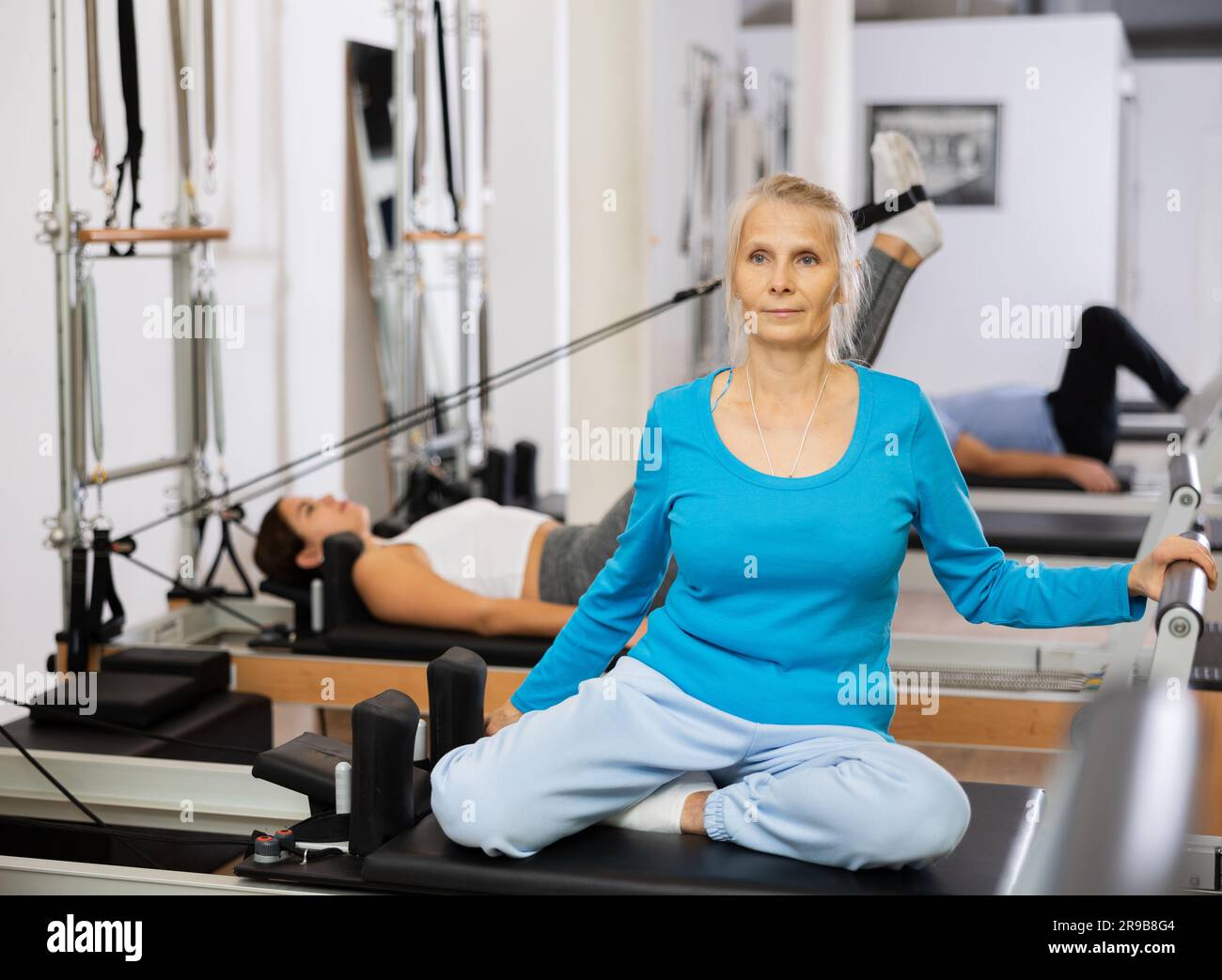 Aged woman in sportive clothes sits with tucked up legs on reformer equipment and training back in rehabilitation center Stock Photo