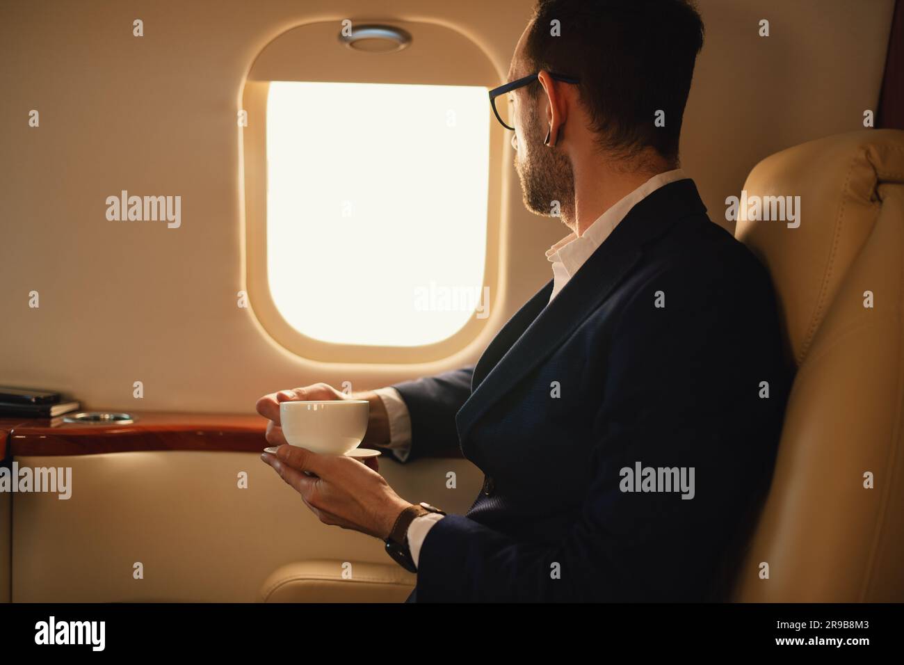 Unrecognizable Elegant CEO Businessman with eyeglasses in a blue jacket sitting inside a private airplane jet drinking coffee looking through a window Stock Photo