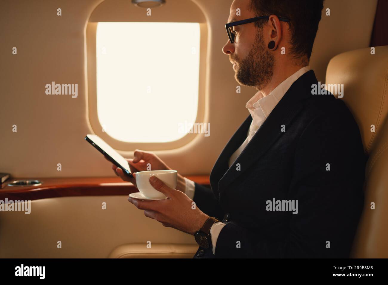 Elegant CEO businessman in eyeglasses and suit sitting inside in first class of a private airplane jet drink of coffee and working with smartphone Stock Photo