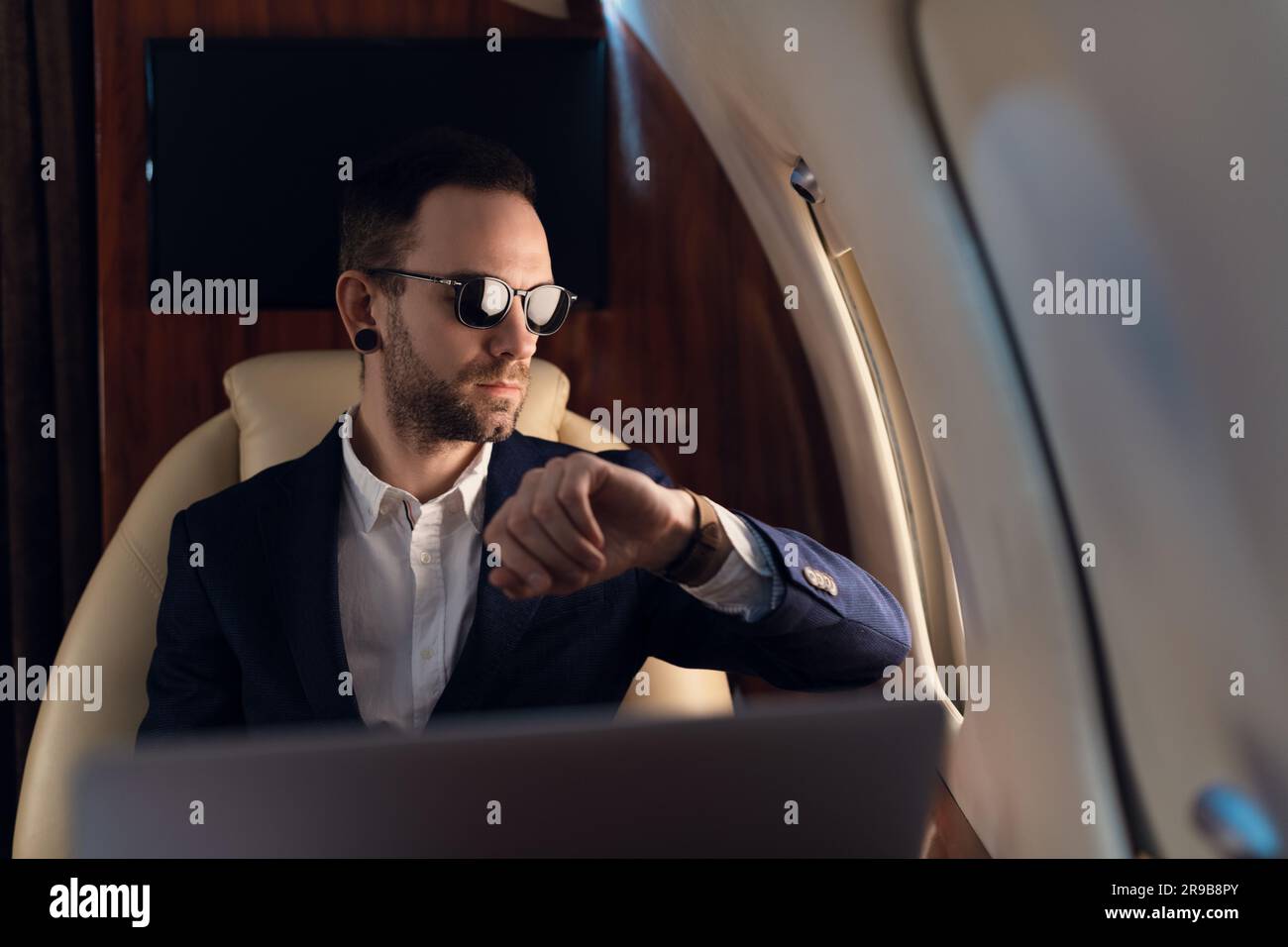 Elegant Confident Businessman with sunglasses in a blue jacket looking through a window in first class corporate business airplane jet Stock Photo