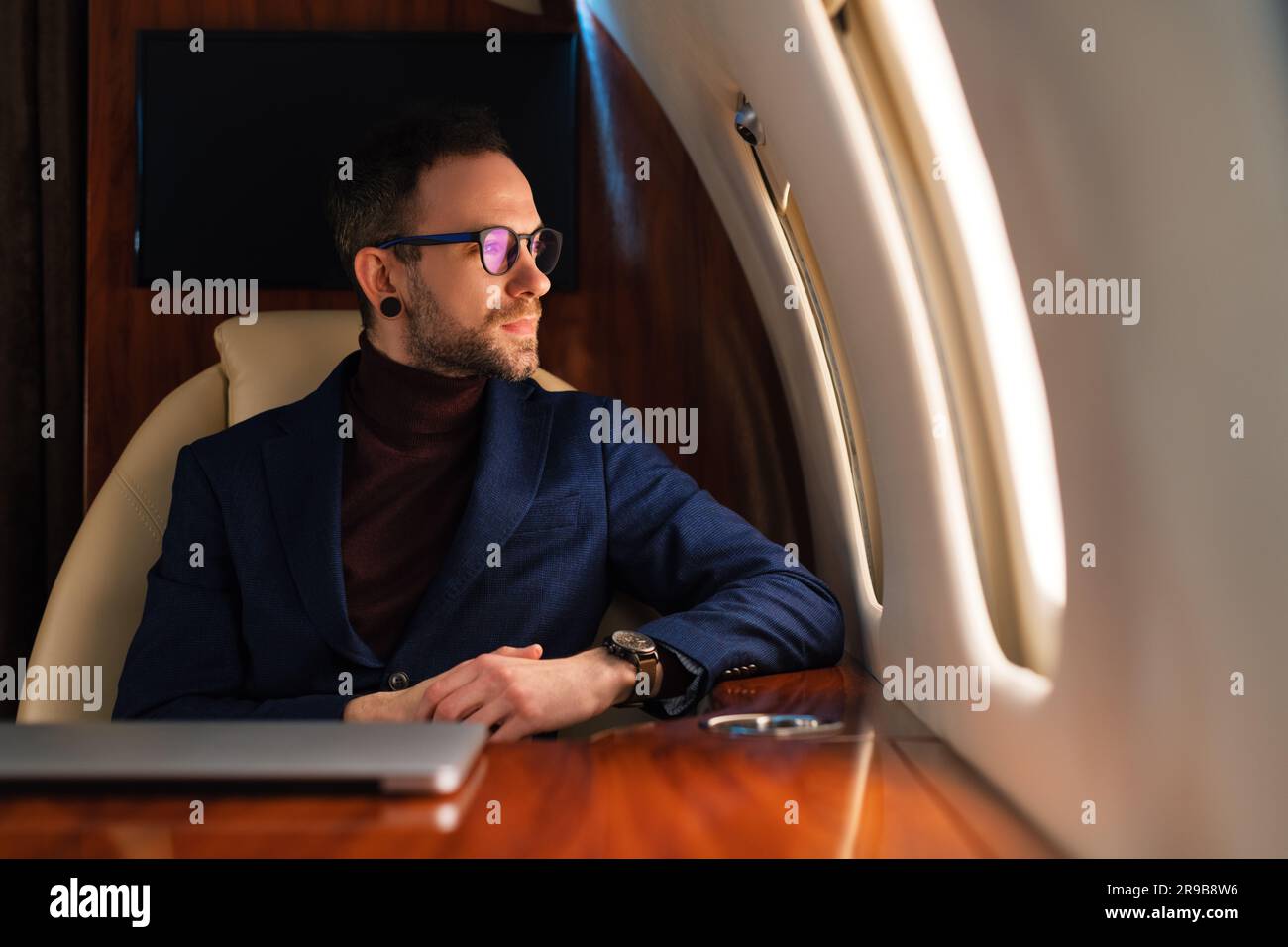 Young Elegant Confident CEO Businessman with eyeglasses in a blue jacket looking through a first-class corporate airplane jet window Stock Photo