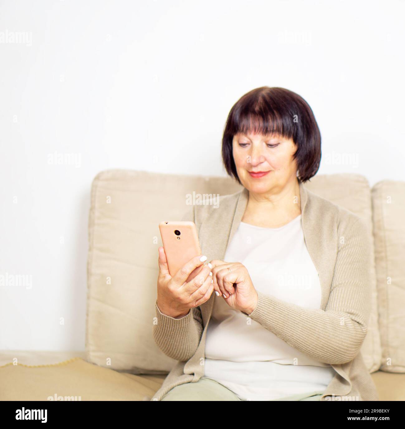 Happy relaxed older woman sitting on comfortable sofa at home and using a mobile phone. Stock Photo
