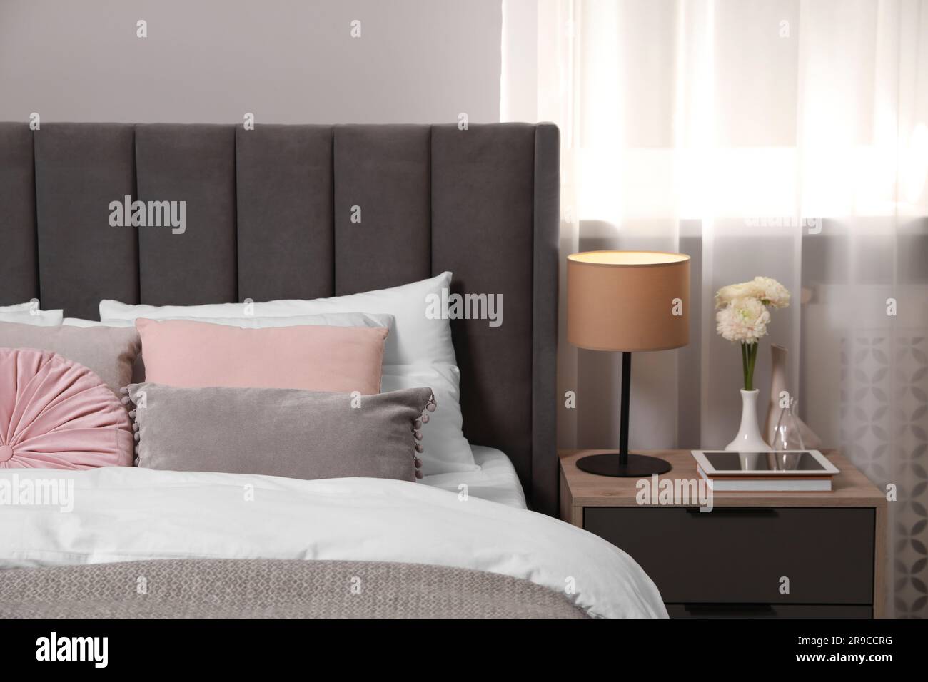 Comfortable bed with cushions, lamp and different decor on bedside table in room. Stylish interior Stock Photo