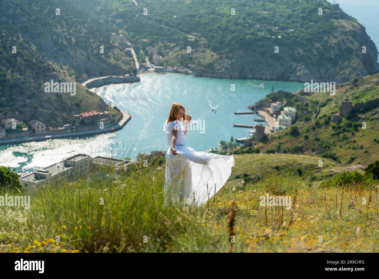 Happy woman in a white dress and hat stands on a rocky cliff above the ...