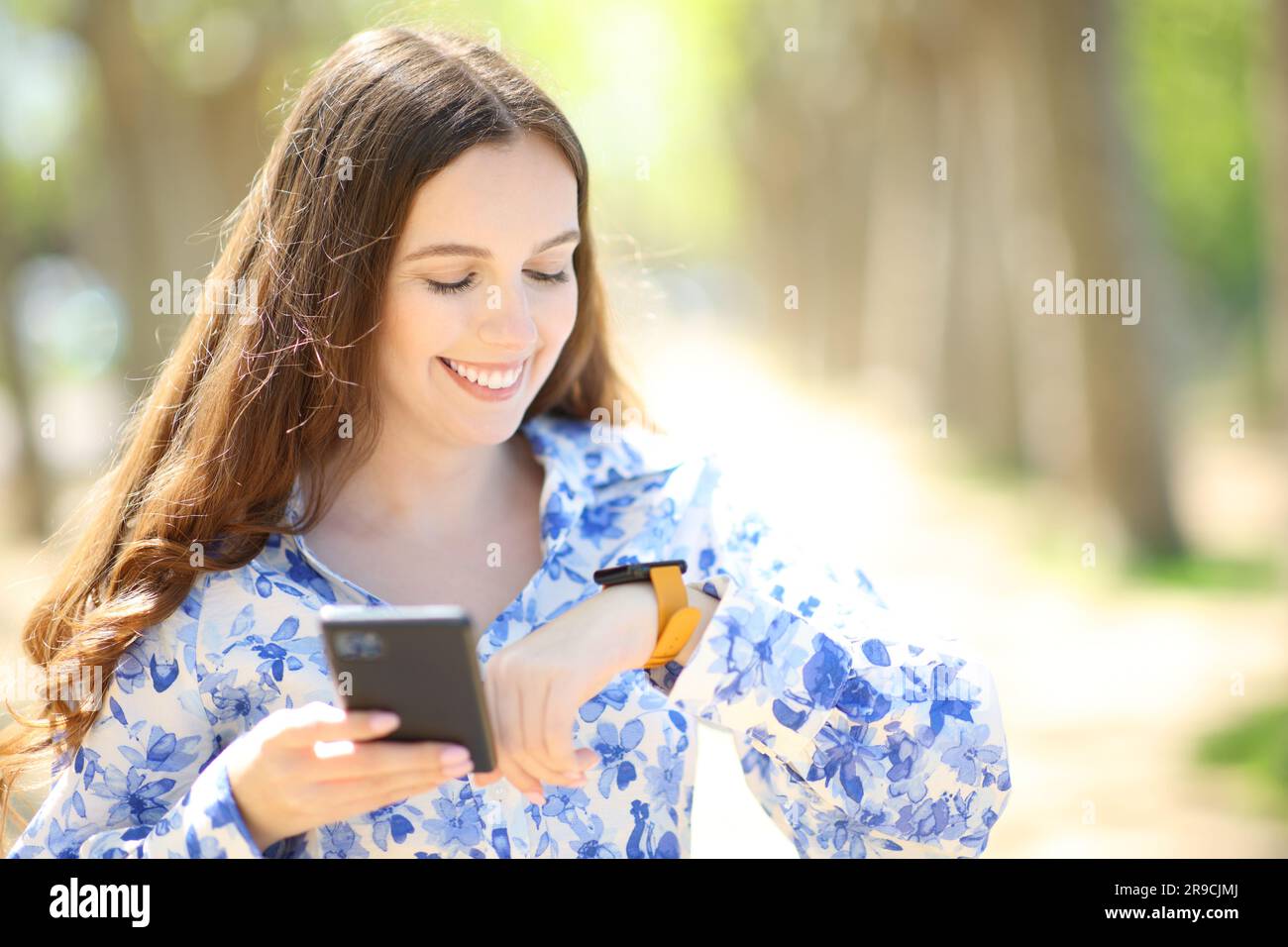 Happy woman holding phone checking smartwatch in a sunny park Stock Photo