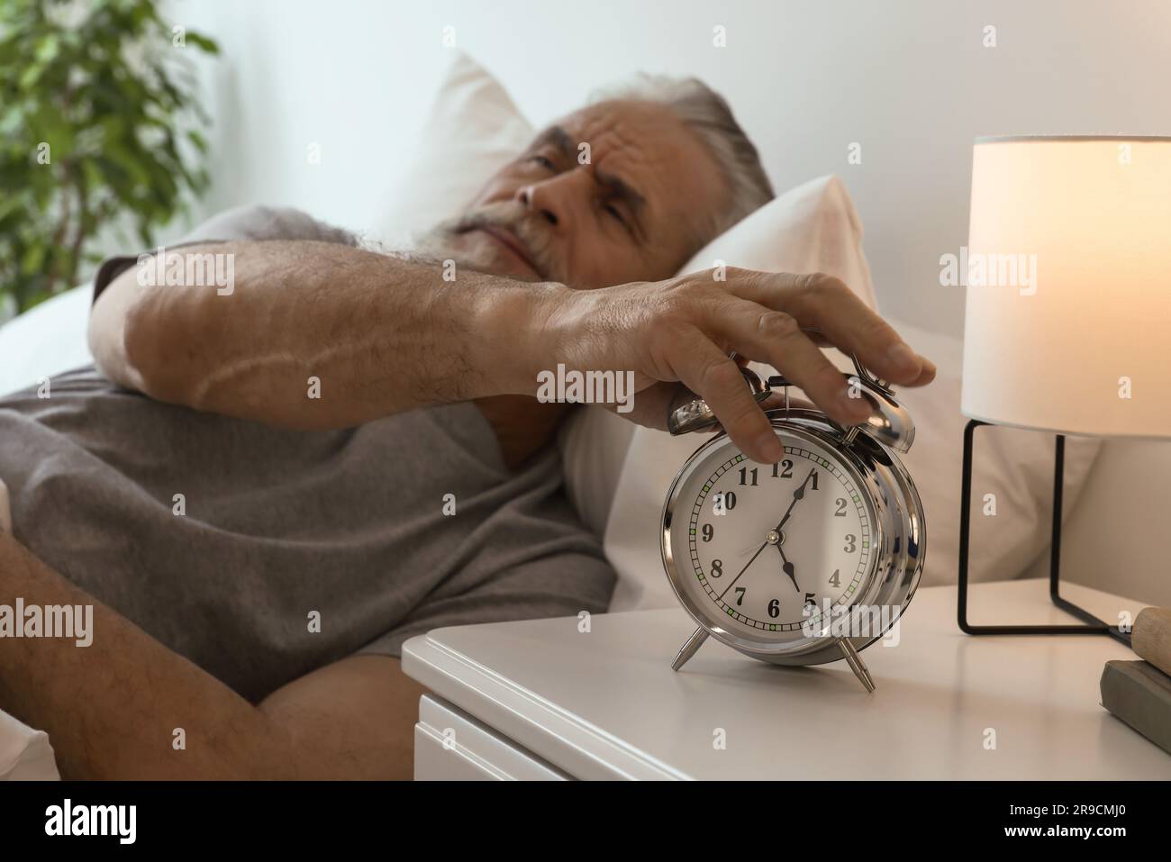 Sleepy senior man turning off alarm clock in bedroom, focus on hand Stock Photo