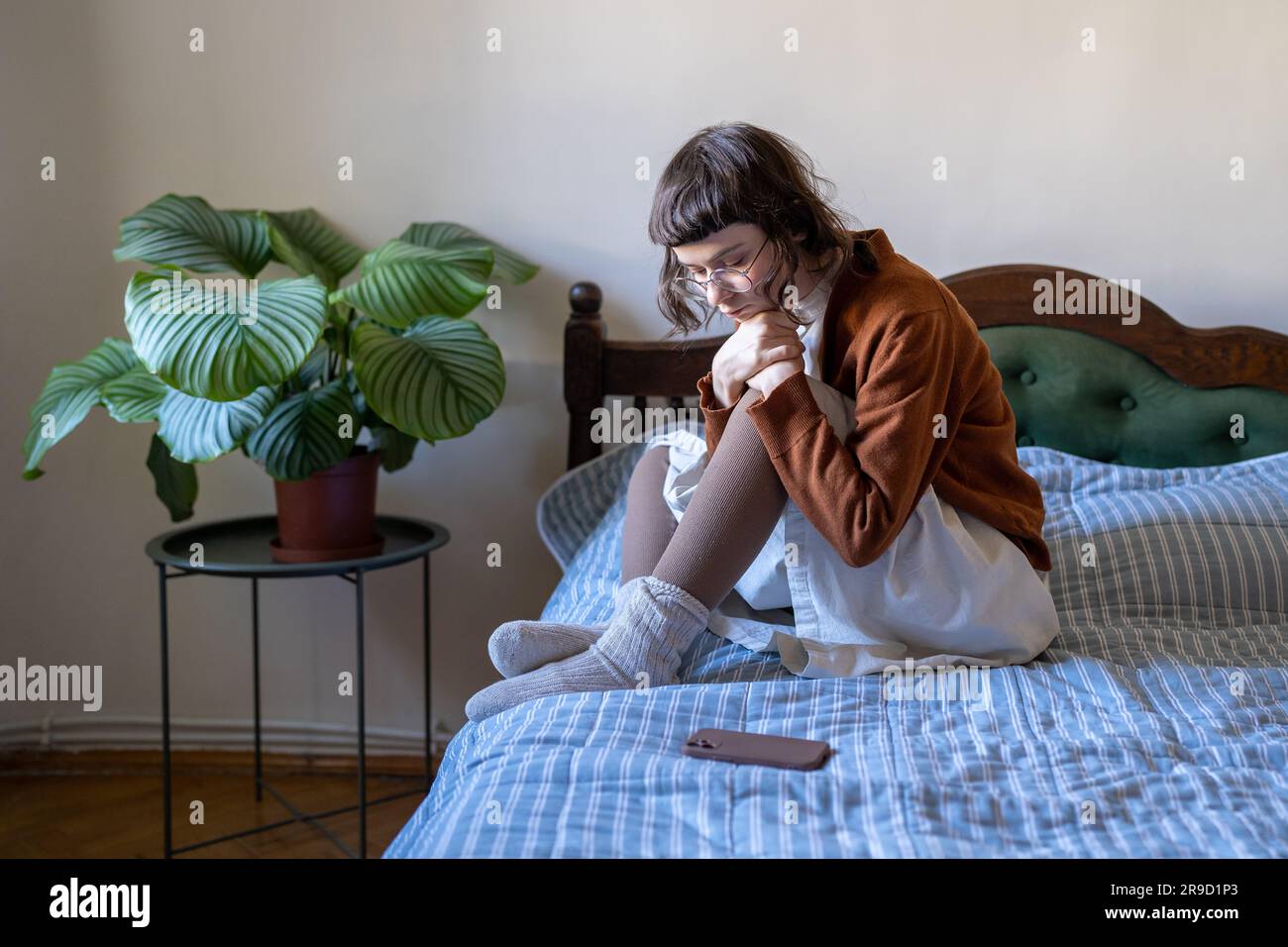 Sad teenage girl in eyeglasses sits on bed, waits for call from boyfriend in difficult relations Stock Photo