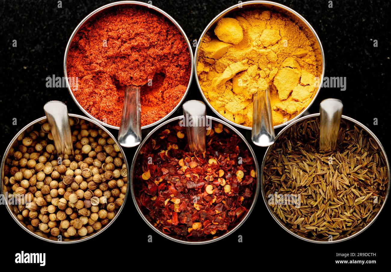 A variety of spices, paprika, turmeric, cumin seeds, crushed chillies, coriander seeds, arranged in a circular formation on a dark background Stock Photo