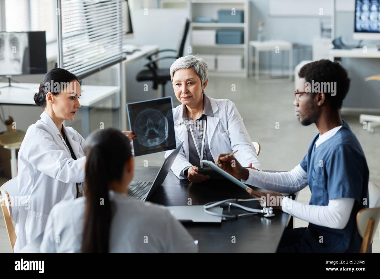 Group of intercultural radiologists discussing scalp x-ray results of patient with traumatic brain injury while sitting by table in medical office Stock Photo