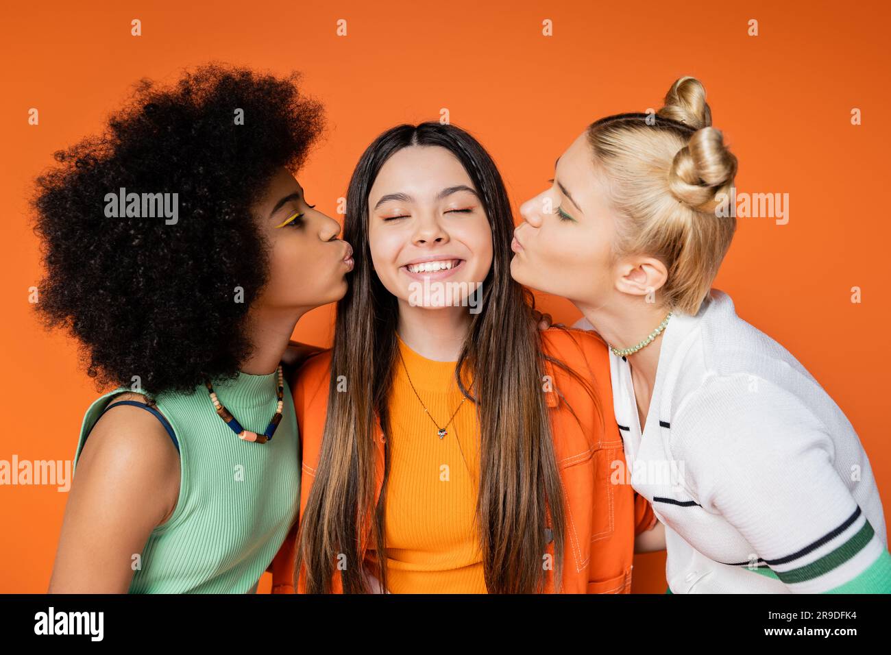 Teen african american and blonde girlfriends with colorful makeup kissing cheerful and stylish girlfriend while standing isolated on orange, trendy ou Stock Photo