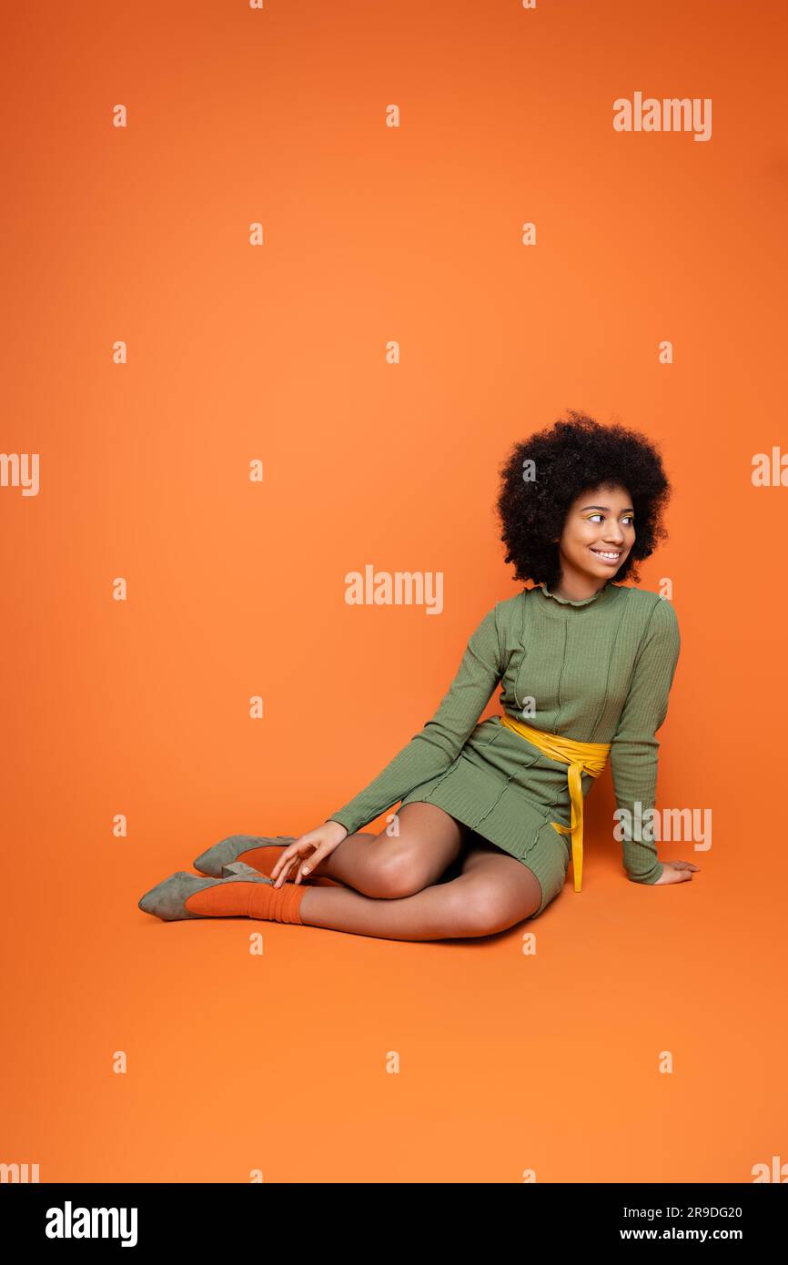 Positive and stylish teen african american girl in green dress looking away while sitting and posing on orange background, youth culture and generatio Stock Photo