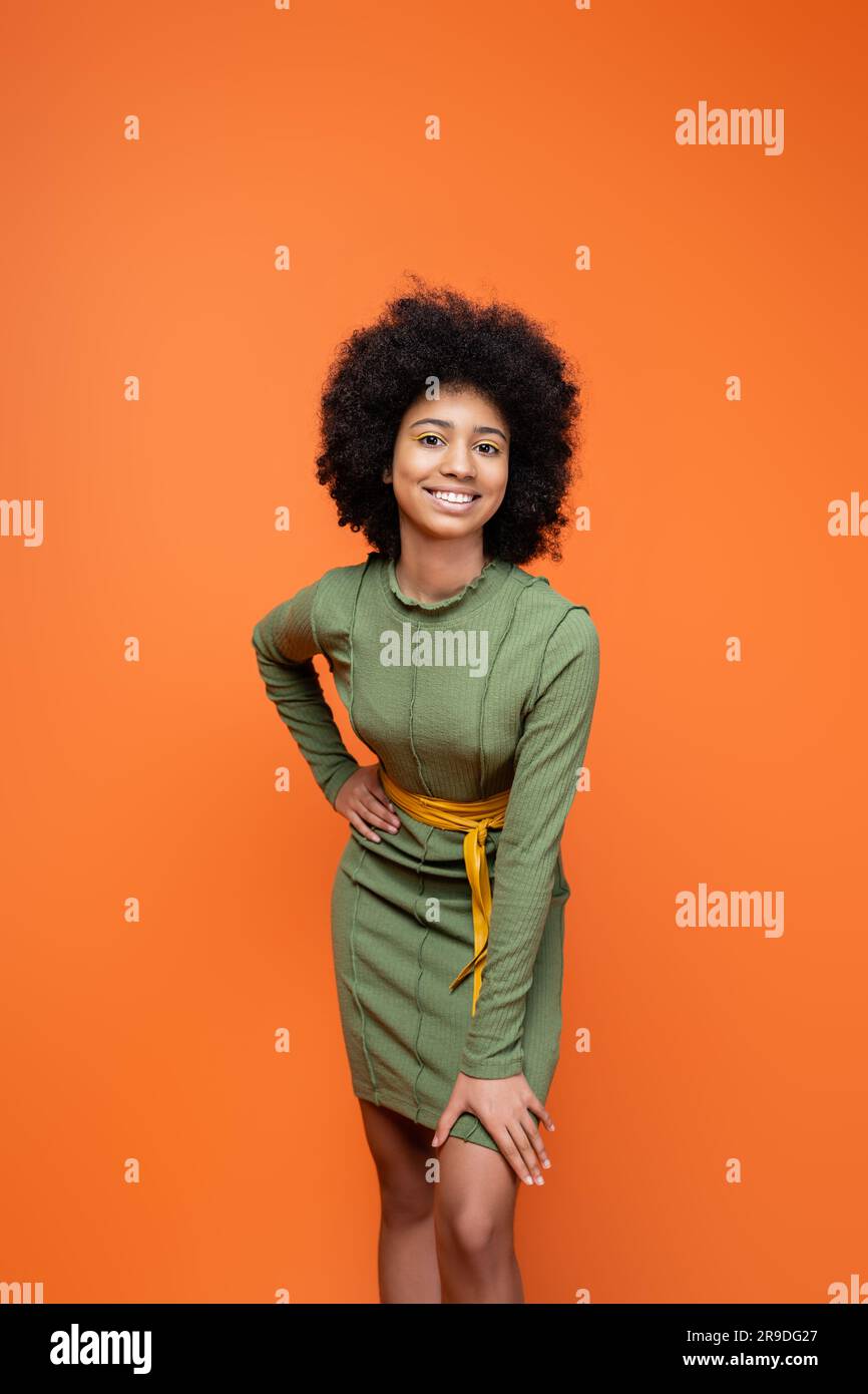 Trendy and positive teenage african american girl with bright makeup wearing green dress and holding hand on hip isolated on orange, youth culture and Stock Photo