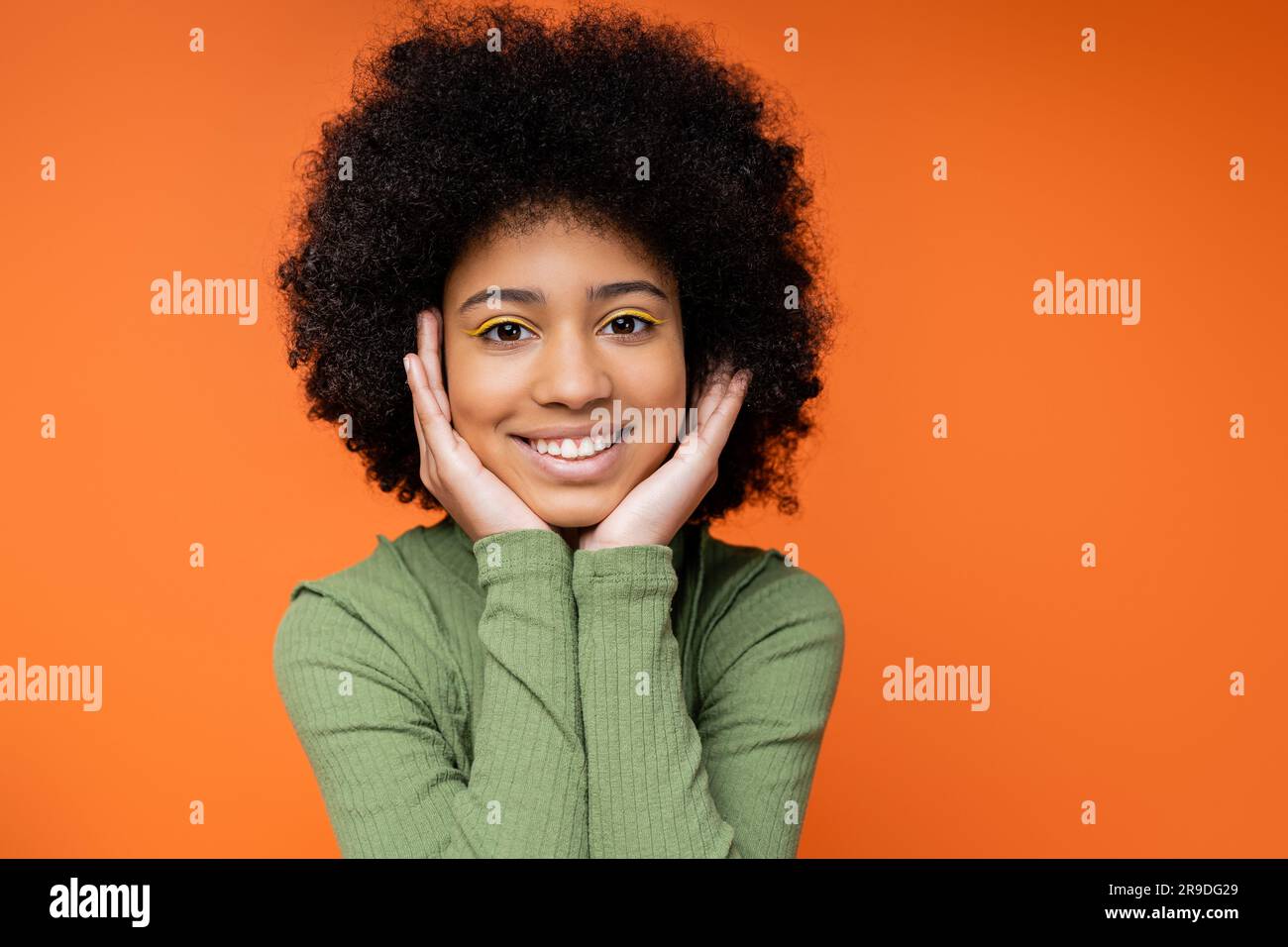 Portrait of happy teen african amerian girl with bold makeup wearing green dress and touching cheeks while standing isolated on orange, youth culture Stock Photo