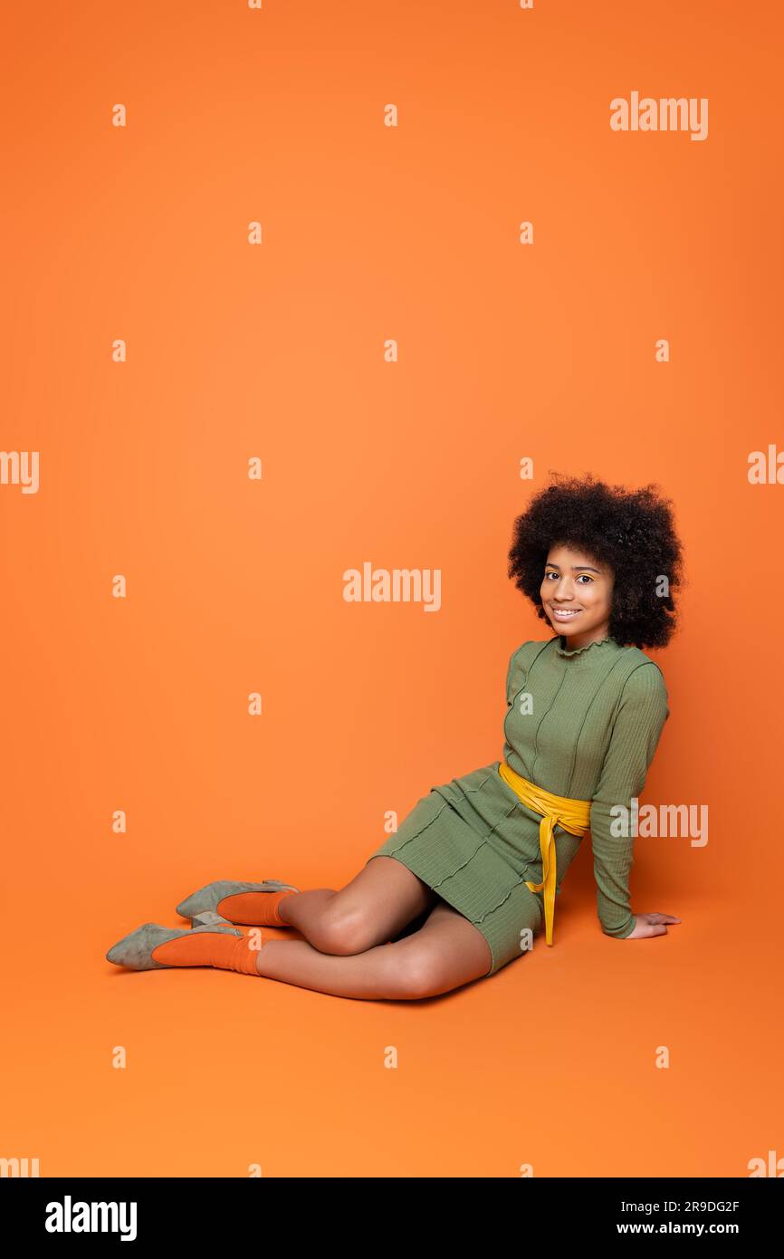 Full length of positive african american teenager with bold makeup and green dress smiling at camera while sitting on orange background, youth culture Stock Photo