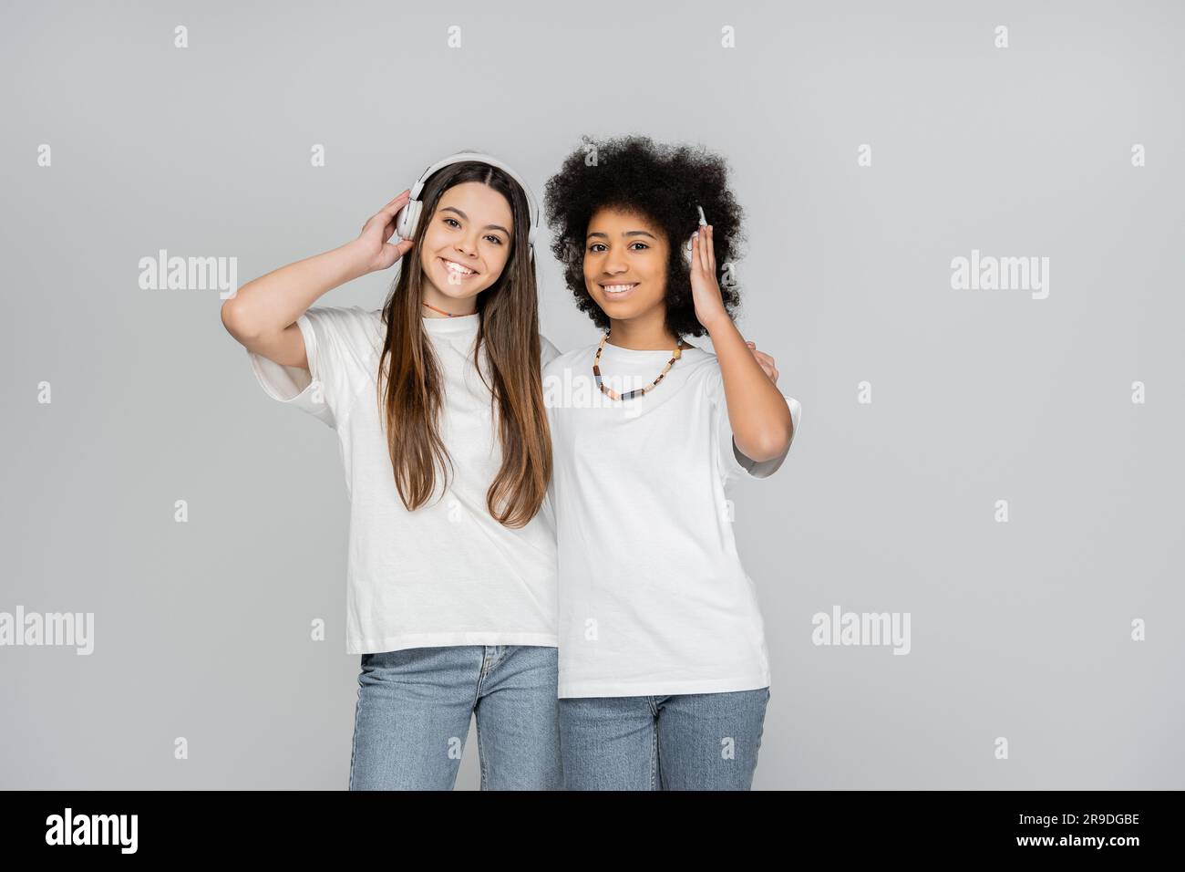 Joyful and interracial teen girls in white t-shirts and jeans listening music in headphones and looking at camera isolated on grey, energetic teenage Stock Photo