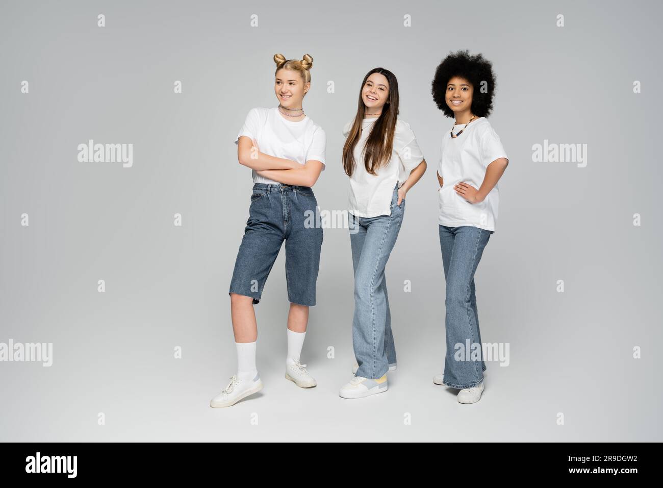 Full length of smiling interracial teen girls in white t-shirts and jeans posing and looking at camera while standing on grey background, lively teena Stock Photo