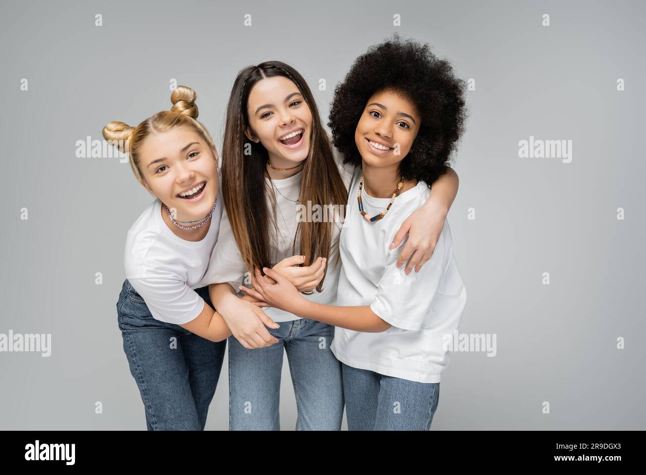 Positive multiethnic teen girls in white t-shirts and jeans hugging brunette friend and looking at camera while standing isolated on grey, lively teen Stock Photo