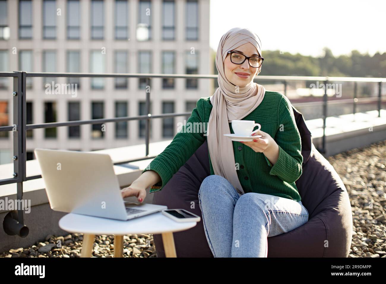 Portrait of young female in hijab drinking coffee while placing laptop ...